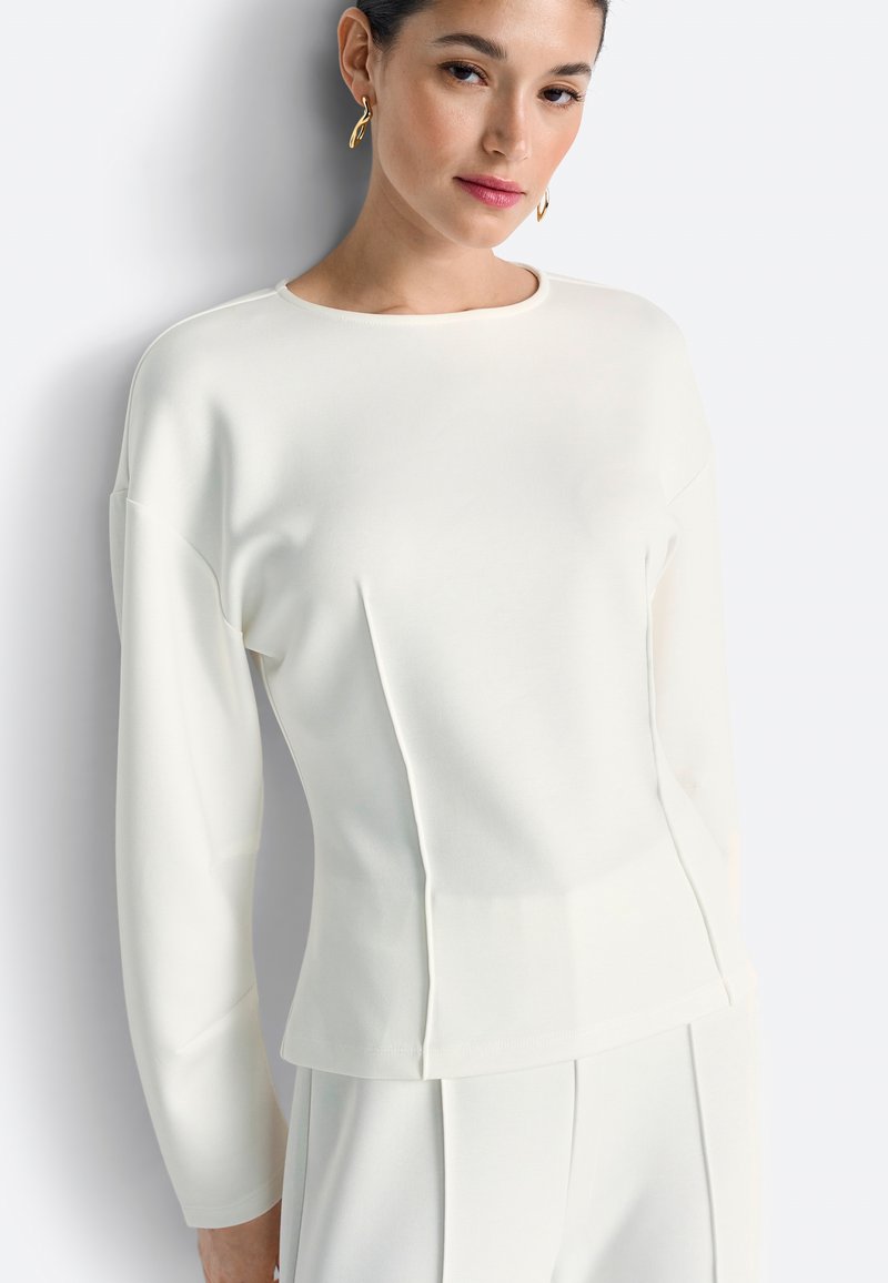 Valge pikkade varrukatega särk ümmarguse kaelusega ja struktureeritud õmblustega, millel on lõikevolditud siluett ja pehme tekstiilipind.