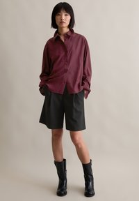 Maroon button-up shirt with a relaxed fit, paired with dark, pleated shorts. Black knee-high boots complete the outfit. Minimalist design.