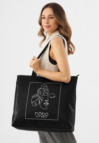 Zwarte draagtas met een minimalistisch ontwerp in witte lijntekeningen van bladeren en een gezicht; vervaardigd uit soepel materiaal met stevige schouderbanden.