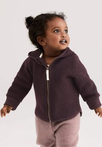 Peuter met krullend haar in een paarse hoodie met rits en roze broek, die naar boven kijkt en glimlacht tegen een effen lichte achtergrond.