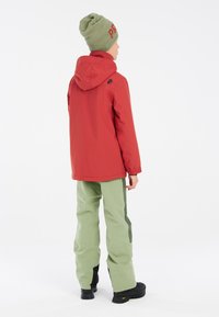 Giacca impermeabile rossa con cappuccio, abbinata a pantaloni impermeabili verdi con pannelli laterali. Stivali da trekking neri completano l'outfit.
