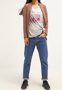 Blazer leggero marrone, t-shirt grafica floreale grigia annodata in vita, jeans blu con orli arrotolati e sneakers nere. Outfit semplice e casual.