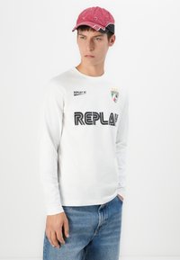 Ung mand iført en rød kasket, hvid langærmet skjorte med teksten "REPLAY" og blå jeans, stående med den ene hånd i lommen.