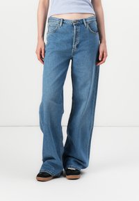 Modello che indossa jeans blu larghi e scarpe nere, in piedi su uno sfondo bianco semplice, con una maglietta a righe parzialmente visibile.