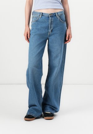 Model in loszittende blauwe jeans en zwarte schoenen, staande tegen een effen witte achtergrond, met een gestreept shirt dat deels zichtbaar is.