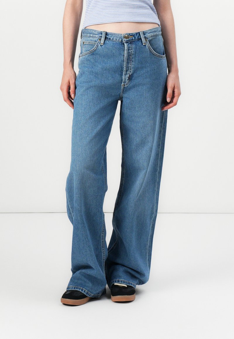 Modello che indossa jeans blu larghi e scarpe nere, in piedi su uno sfondo bianco semplice, con una maglietta a righe parzialmente visibile.