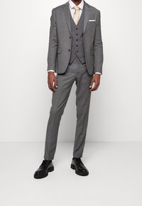 Szary garnitur trzyczęściowy z klapami typu notch, detalami w postaci guzików oraz spodniami dopasowanymi. Połączony z beżowym krawatem i błyszczącymi czarnymi butami.
