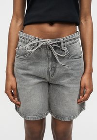 Γυναίκα που φορά ψηλόμεσο γκρι σορτς τζιν με ζώνη για δέσιμο και μπροστινές τσέπες, σε συνδυασμό με ένα μαύρο κοντό αμάνικο τοπ.