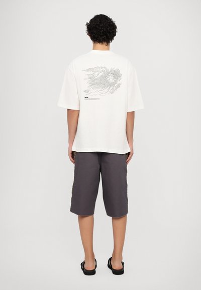 Witte katoenen t-shirt met een graphic op de achterkant; gecombineerd met grijze shorts met zijaccenten. Model draagt zwarte sandalen.