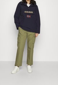 Marine kleur pullover jacket met een voorzak, logo detail en vlag patch; gecombineerd met olijfgroene cargo broeken en witte sneakers.