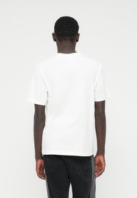 Λευκό βαμβακερό μπλουζάκι με κοντά μανίκια, στρογγυλή λαιμόκοψη και άνετη γραμμή· εμφανίζεται από πίσω, συνδυασμένο με σκούρο τζιν.