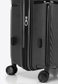 Valise rigide noire avec une surface texturée, double fermeture éclair et deux roulettes faciles à manœuvrer.