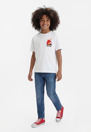 Nasmejan otrok s kodrastimi lasmi, oblečen v belo Moschino majico, modre kavbojke in rdeče superge, stoji pred belim ozadjem.