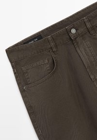 Gros plan sur un pantalon marron foncé montrant la poche avant, les passants de ceinture et la fermeture par bouton avec une étiquette de marque visible à l'intérieur de la ceinture.