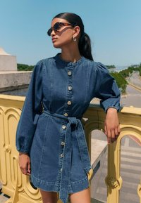 Denimkleit, millel on puff-varrukad, nööpidega esikülg, vöö läbi seotud ja kuldsete detailidega aktsendid. Modell kannab kilpkonnakujulisi päikeseprille, taustal on sinine taevas.