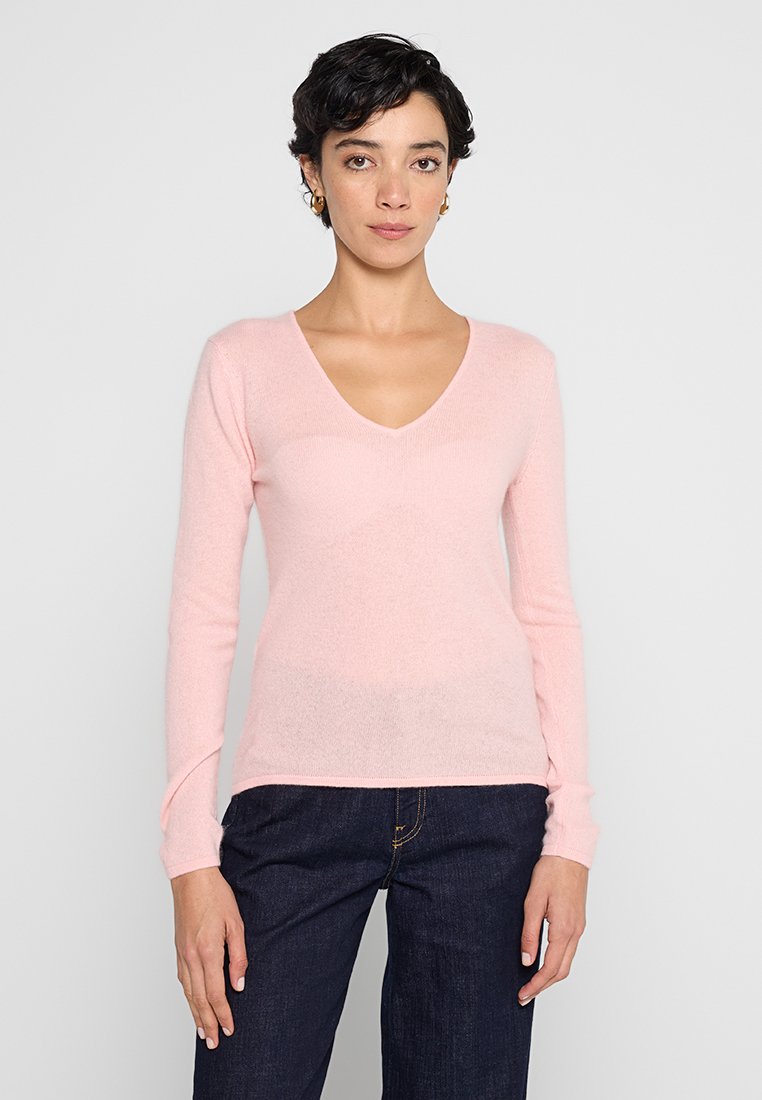 Jasnoróżowy sweter dzianinowy z dekoltem w serek, długimi rękawami i miękką fakturą. Noszony z ciemnoniebieskimi dżinsami. Idealny na codzienne wyjścia.