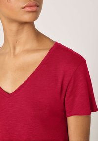 Raudonas V-apykaklės marškinėlis, pagamintas iš minkštos, tekstūruotos medžiagos. Turi trumpas rankoves ir laisvą siluetą, o apykaklės siuvinėjimas yra subtilus.