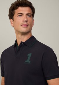 Námornícky polo tričko s zelenými akcentmi na golieri a vyšívaným logom "1" a "HACKETT" na hrudi, so klasickým strihom a hladkou textúrou.