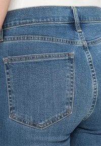 Un jean en denim bleu présente une poche arrière droite avec des coutures contrastées. Le tissu a une texture légèrement usée avec une finition de lavage moyen.