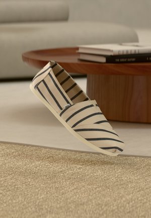 Beige canvas instapschoen met zwarte horizontale strepen zwevend boven een geweven tapijt in een woonkamer met een ronde houten tafel en boeken.