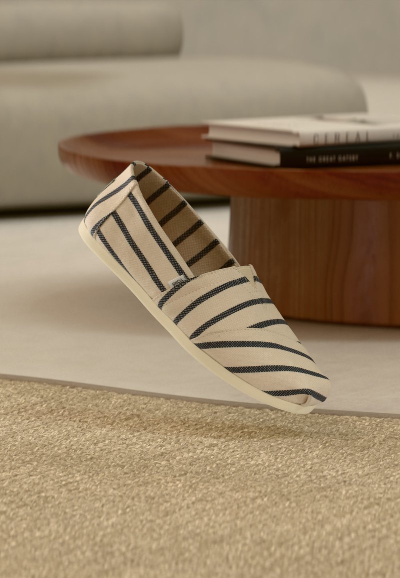 Μπεζ καμβάς παπούτσι χωρίς κορδόνια με μαύρες οριζόντιες ρίγες που αιωρείται πάνω από ένα υφαντό χαλί σε ένα σαλόνι με στρογγυλό ξύλινο τραπέζι και βιβλία.