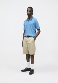 Lys blå teksturert poloshirt med krage, kombinert med beige shorts og svarte sko. Modellen står med hendene i lommene, og hvite sokker er synlige.
