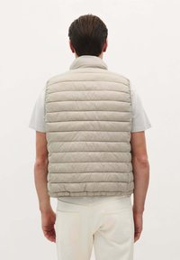Polstret ærmeløs vest i lys beige, med vertikalt quiltet design, høj krave og en glat tekstur. Bærer den over en hvid skjorte.