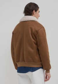 Braune Wildleder-Bomberjacke mit einem flauschigen weißen Pelzkragen, gerippten Bündchen und Saum. Sie hat einen lockeren Schnitt und eine glatte Textur.