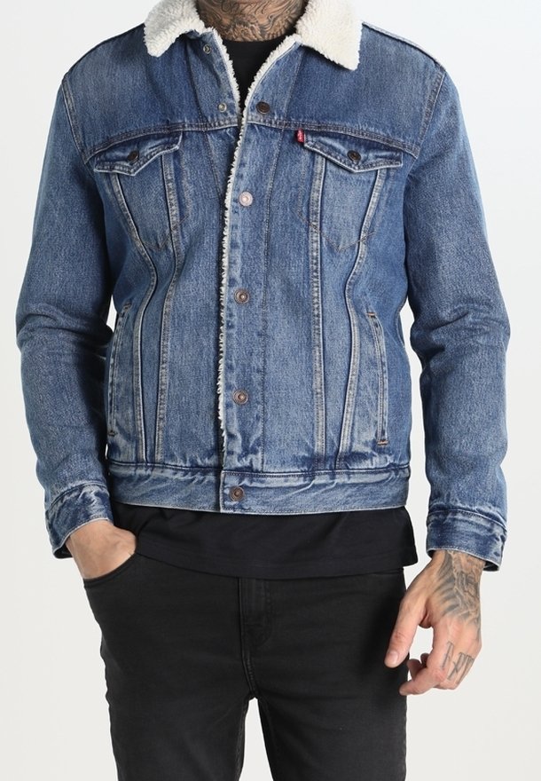 Homme portant une veste en jean bleue avec un col en polaire blanche, une chemise noire en dessous, et un pantalon noir, avec une main dans la poche et des tatouages visibles sur la main.