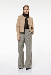 Beżowa strukturalna kurtka z czarnymi wykończeniami, zapinana na guziki, noszona na brązowej bluzce. Szare szerokie spodnie w kratę sięgają do podłogi.