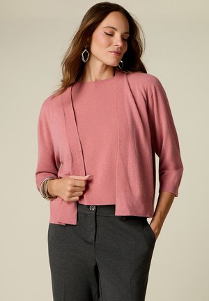 Donna con lunghi capelli castani che indossa un cardigan rosa e una maglietta, pantaloni grigio scuro, orecchini geometrici e braccialetti, posando con una mano in tasca.