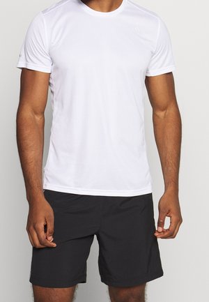 Mand iført en almindelig hvid kortærmet sportstrøje og sorte knælange shorts, står mod en neutral baggrund.