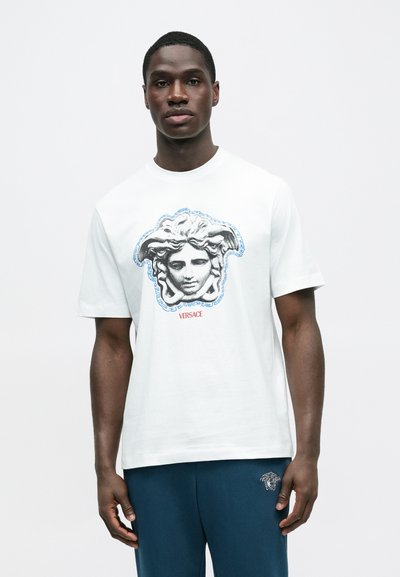 Λευκό βαμβακερό T-shirt με κεντρική εικόνα ενός ελληνικού μυθολογικού κεφαλιού, περιτριγυρισμένο από μπλε λεπτομέρειες και τη λέξη "VERSACE" με κόκκινα γράμματα.