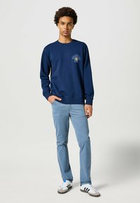 Sudadera navy de manga larga y cuello redondo, con un logo gráfico en el pecho, combinada con jeans de un azul claro y zapatillas deportivas.