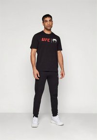 Must UFC kaubamärgiga t-särk, millel on punane logo rinnal, kombineeritud mustade dressipükste ja valgete tossudega, kõik mõõdukas tänava stiilis.