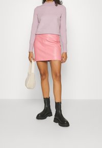 Pull ajusté violet clair à manches longues, associé à une jupe courte en cuir rose brillant. Bottines noires à semelles épaisses ; tenant un sac beige.