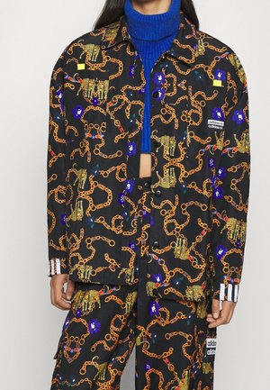 Veste et pantalon noirs avec des motifs chaîne orange, léopard et fleurs bleues, portés sur un pull à col roulé bleu.