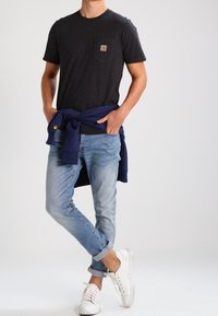 T-shirt nera in cotone con taschino sul petto, abbinata a jeans in denim blu chiaro. Sneakers bianche e un maglione blu navy legato in vita.