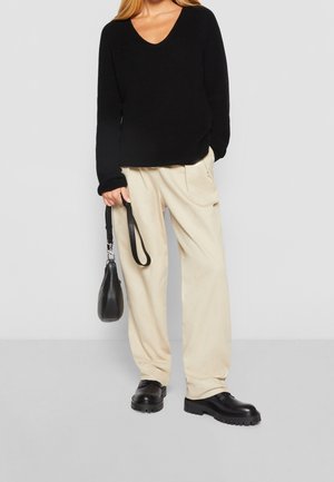 Vrouw met een zwarte V-hals trui, beige broek met wijde pijpen, zwarte chunky schoenen, en een klein zwart handtasje aan haar zijde.