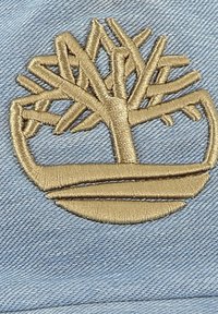 Logo ad albero ricamato beige su tessuto azzurro chiaro con una texture intrecciata, che rappresenta rami e una base curvata.