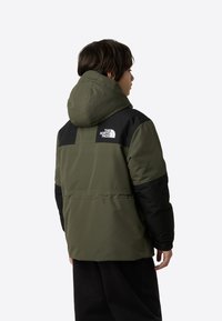 Grønn og svart isolert jakke med hette, med en tydelig logo på ryggen og et strukturert design med middels lengde.