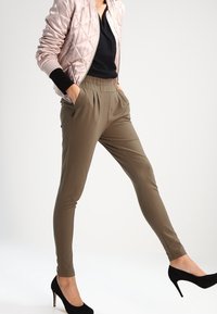 Chaqueta bomber acolchada rosa claro, blusa negra, pantalones tapered verdes olivo y tacones altos negros. La chaqueta tiene puños de canalé y cuello alzado.
