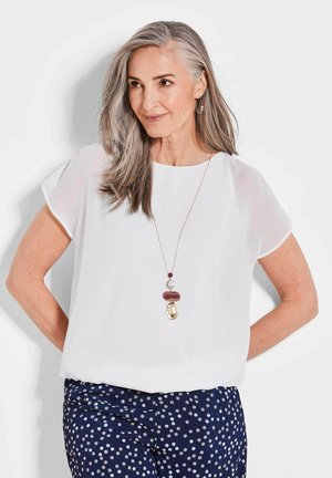 Blouse blanche à manches courtes en tissu léger, associée à un pantalon à pois bleu marine. Présente un collier en pendentif avec plusieurs pierres de couleurs.