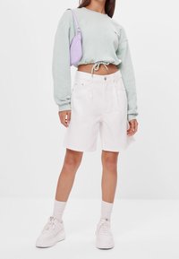 Světl zelený zkrácený svetr s vázáním v pase, kombinovaný s bílými džínovými kraťasy. Doplňky zahrnují malou levandulovou kabelku a bílé tenisky.