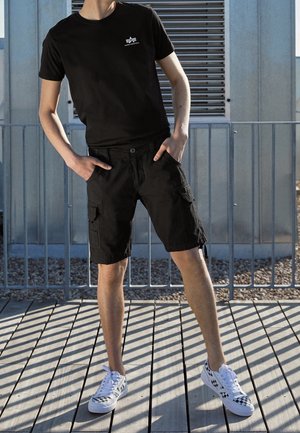 Person iført sort t-shirt og cargobukser står på en træterrasse med hænderne i lommerne og har hvide sneakers med sorte mønstre på.