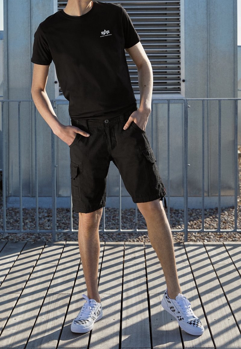 Personne portant un t-shirt noir et un short cargo se tient sur une terrasse en bois, les mains dans les poches, chaussée de baskets blanches à motifs noirs.