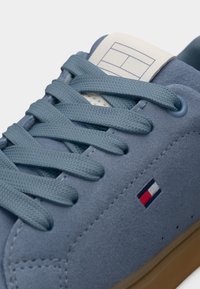 Ανοιχτό μπλε σουέντ αθλητικό παπούτσι με επίπεδα κορδόνια, αντίθετη μπεζ σόλα και ένα μικρό πολύχρωμο έμβλημα στο πλάι κοντά στα κορδόνια.