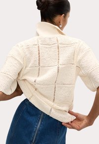 Haut en tricot couleur crème avec un col montant, présentant un motif de mailles ouvertes et des manches courtes, associé à une jupe en denim foncé.