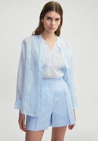 Helles blaues Leinenhemd, durchsichtig mit einer geknöpften Vorderseite, kombiniert mit hellblauen maßgeschneiderten Shorts. Ausgefallenes Spitzendetail im Inneren des Blusens.