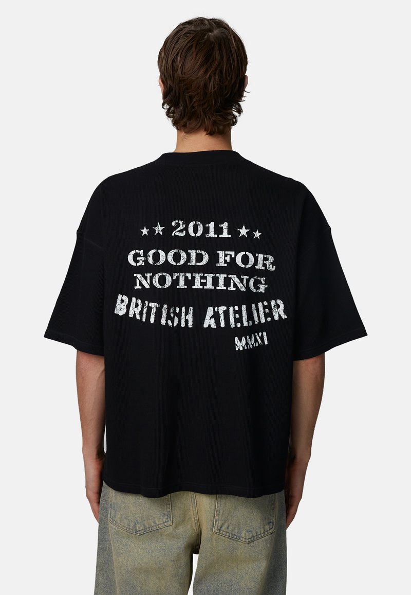 Mężczyzna z krótkimi brązowymi włosami, ubrany w czarny, oversize'owy T-shirt z białym napisem „2011 Good For Nothing British Atelier MMXI” na plecach.